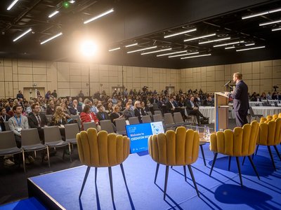 Großer Saal mit vielen Leuten; Bühne; ein Mann präsentiert im Vordergrund 