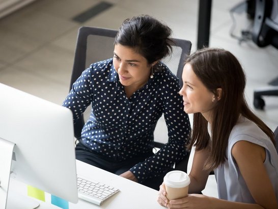 Multiracial colleagues indian and caucasian young women having coffee break sitting together at desk in office. Diverse students interns friends looks at pc screen talking discussing working moments