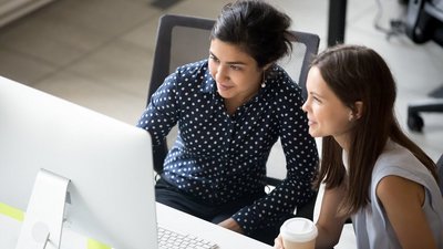 Multiracial colleagues indian and caucasian young women having coffee break sitting together at desk in office. Diverse students interns friends looks at pc screen talking discussing working moments