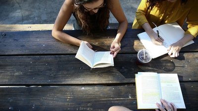 Eine Gruppe von Menschen sitzt an einem rustikalen Holztisch im Freien. Eine Person liest ein Buch, eine andere schreibt in ein Notizbuch, und auf dem Tisch liegt ein weiteres geöffnetes Buch mit Textmarkierungen. Ein Plastikbecher mit Getränk steht neben den Büchern.