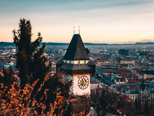 Eine atemberaubende Luftaufnahme von Graz mit dem ikonischen Uhrturm, umgeben von üppigem Laubwerk in der Dämmerung.