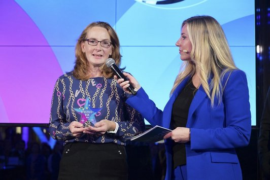 Zwei Frauen stehen auf der Bühne, eine Frau hält das Mikrofon, die andere Dame, welche einen Pokal in der Hand hält, spricht
