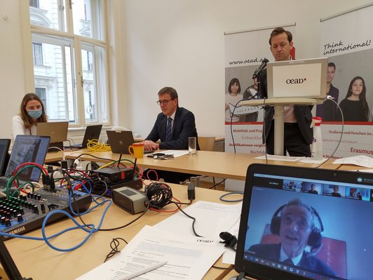 Table with three speakers, two in front of it sitting in front of a laptop, one standing at the desk, and in the right foreground a screen with an external speaker connected