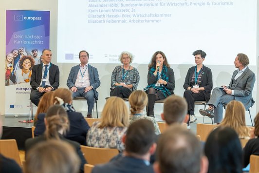 Die 6 Panelisten der Diskussion sitzen auf der Bühne, hinter ihnen auf der Bühne ist die Präsentation eingeblendet mit der Übersicht ihrer Namen. 