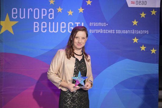 Frau in schwarzem Kleid und beigem Blazer hält eine Auszeichnung (Stern) in der Hand.