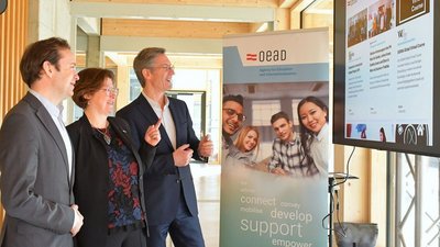 Zwei Männer und eine Frau stehen zusammen und richten ihren Blick interessiert auf einen großen Bildschirm