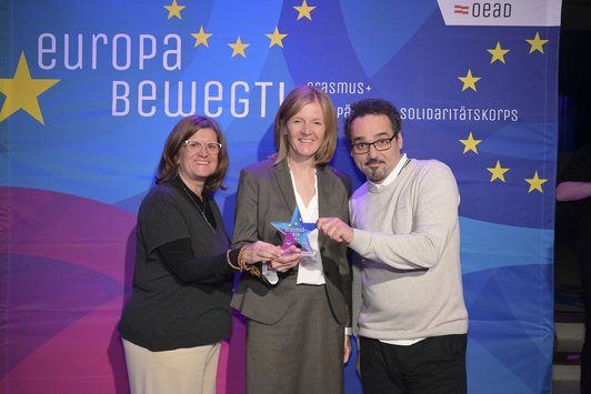 Zwei Frauen und ein Mann stehen vor einem Transparent mit der Aufschrift "Europa bewegt" und lachen in die Kamera.