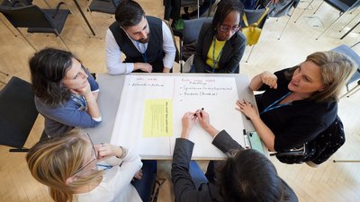 Gruppe von Personen sitzt um einen Tisch, vor ihnen ein Flipchart-Papier und ist in gemeinsames Gespräch vertieft