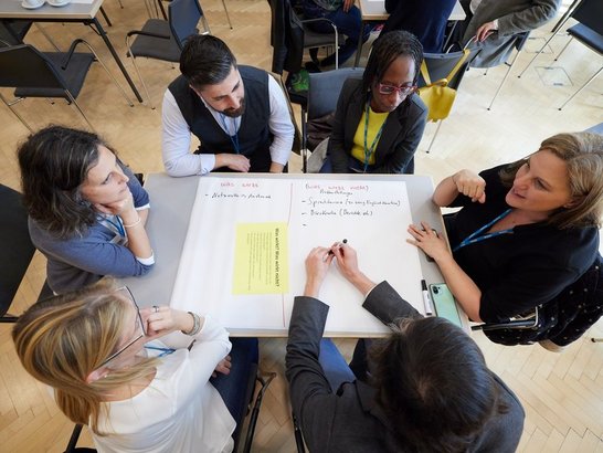 Gruppe von Personen sitzt um einen Tisch, vor ihnen ein Flipchart-Papier und ist in gemeinsames Gespräch vertieft