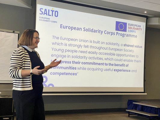 Dame präsentiert mit ausgestreckten Armen vor einer Leinwand mit der Aufschrift "European Solidarity Corps Programme"