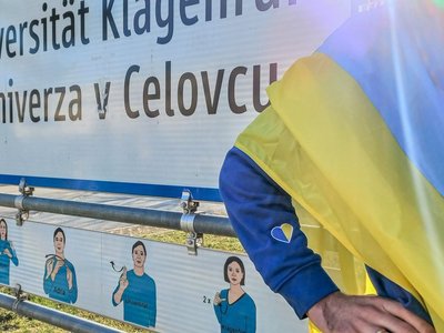 Eine Person mit ukrainischer Flagge steht neben einem Ortsschild mit dem Text "Universität Klagenfurt/Univerza v Celuvcu"