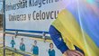 Eine Person mit ukrainischer Flagge steht neben einem Ortsschild mit dem Text "Universität Klagenfurt/Univerza v Celuvcu"