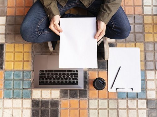 Jemand sitzt im Schneidersitz am boden und hält ein Blatt Papier in den Händen. Vor im stehen ein Laptop, ein Kaffebecher, Papier und ein Stift.