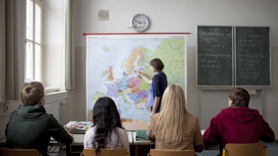 Vier Schüler und Schülerinnen blicken auf die Tafel, dort steht eine Lehrerin an einer Weltkarte