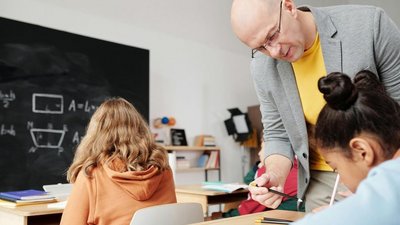 Mann im grauen Pullover (Lehrer) mit Bleistift in der Hand neigt sich zu einer Schülerin