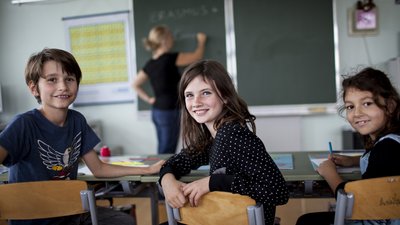 Drei sitzende Volksschulkinder in Klassenraum drehen sich von Tafel weg und blicken in die Kamera