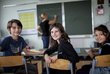 Drei sitzende Volksschulkinder in Klassenraum drehen sich von Tafel weg und blicken in die Kamera