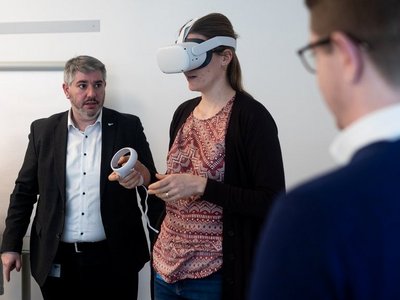 Eine Frau trägt eine VR-Brille und bedient eine VR-Steuerung. Zwei Männer schauen ihr dabei zu.