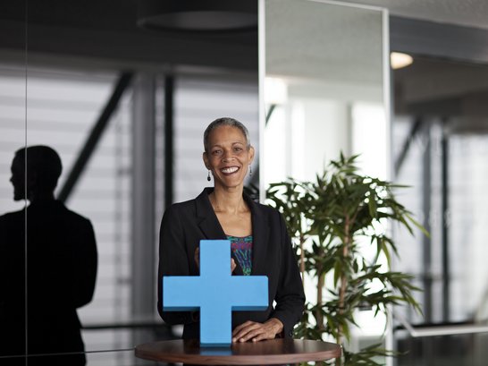 Frau hält ein blaues Erasmus+ Symbol in die Kamera. Hinter ihr steht ein Strauch. 