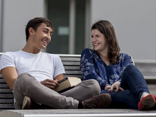 Ein junger Mann und eine junge Frau sitzen auf einer Bank und lachen.